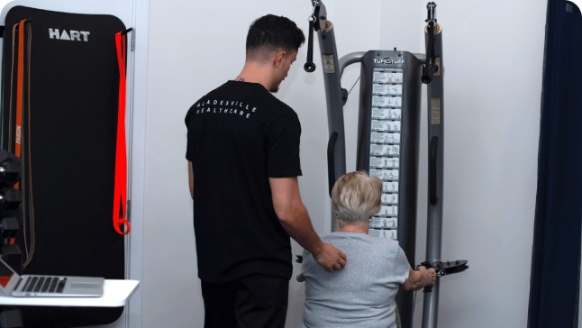 A man assists an older woman using a weight machine in a clinical exercise room.