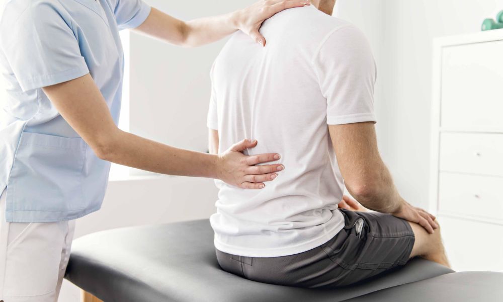Physio for back pain: Healthcare professional examines a seated man's back in a medical office.